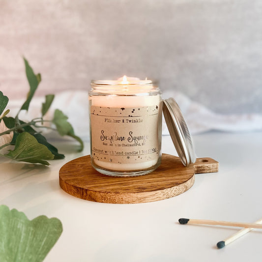 A lit "Sunshine Squeeze" candle by Flicker And Twinkle, handmade with coconut wax, sits on a wooden coaster. The glass jar’s lid leans against it. Matches and green leaves are placed nearby, all on a white surface with a light, neutral background.