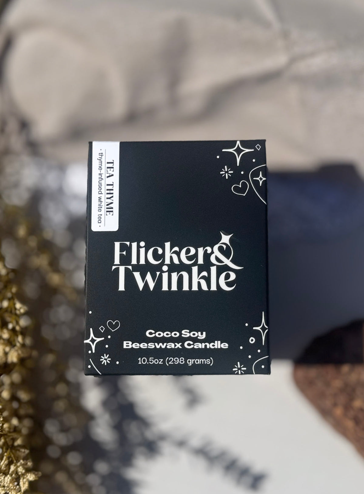 A square black candle box labeled 'Flicker & Twinkle' with white decorative text and star motifs, photographed on a softly blurred surface with warm and cool lighting.