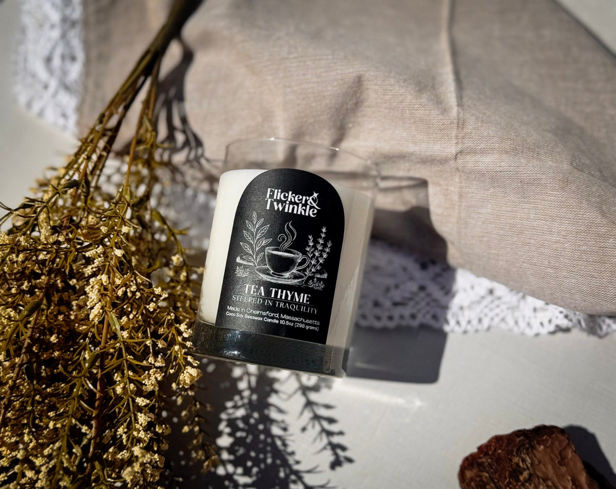 A tea thyme scented candle by Flicker & Twinkle in a clear glass jar with a black label, styled on a white surface with dried flowers, a beige fabric cushion, and a white lace doily.