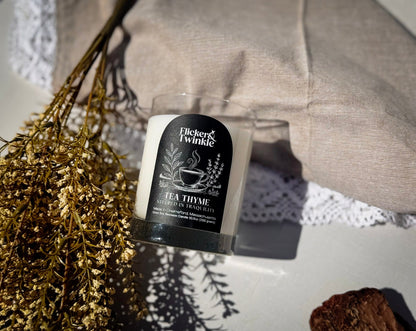 A tea thyme scented candle by Flicker & Twinkle in a clear glass jar with a black label, styled on a white surface with dried flowers, a beige fabric cushion, and a white lace doily.