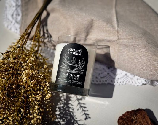 A tea thyme scented candle by Flicker & Twinkle in a clear glass jar with a black label, styled on a white surface with dried flowers, a beige fabric cushion, and a white lace doily.