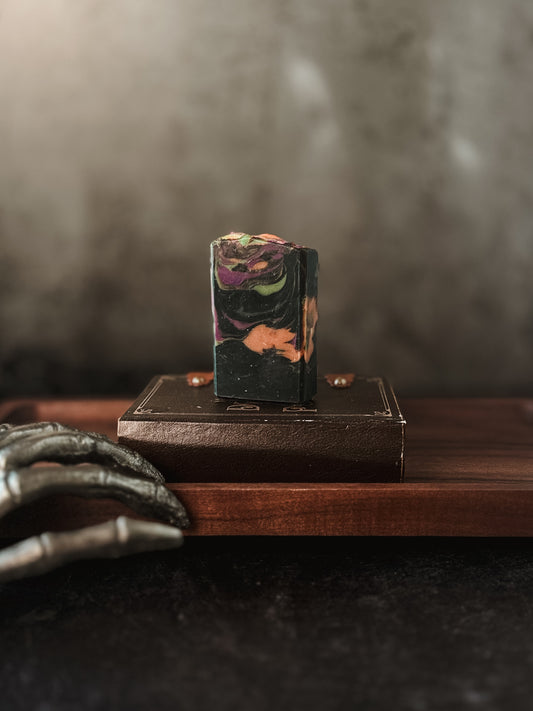 A bar of Flicker And Twinkle's That Witch Goat Milk Soap, featuring a marbled design in dark colors—black, green, purple, and orange—rests on a rectangular wooden tray. A skeletal hand appears in the lower-left corner, adding a spooky touch to the scene. The background is blurred and dark.