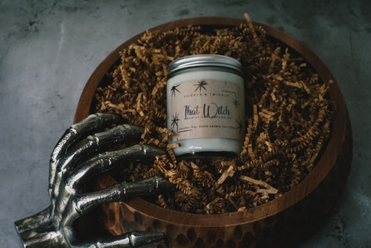 A white Flicker And Twinkle "That Witch" Coconut Wax Candle sits in a glass jar within a wooden bowl filled with shredded brown paper, exuding a spell-binding scent. A metallic skeletal hand leans against the side of the bowl. The background features a textured gray surface, enhancing the candle's enchanting aura.