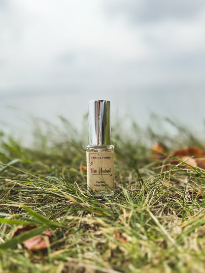 A small glass bottle with a silver cap, containing a yellowish liquid, is placed on grass with a few scattered brown leaves. The label on the bottle features "The Naiad" and some other text under the brand name "Flicker And Twinkle." The background shows a blurry sky, giving a serene outdoor setting that exudes coastal elegance.