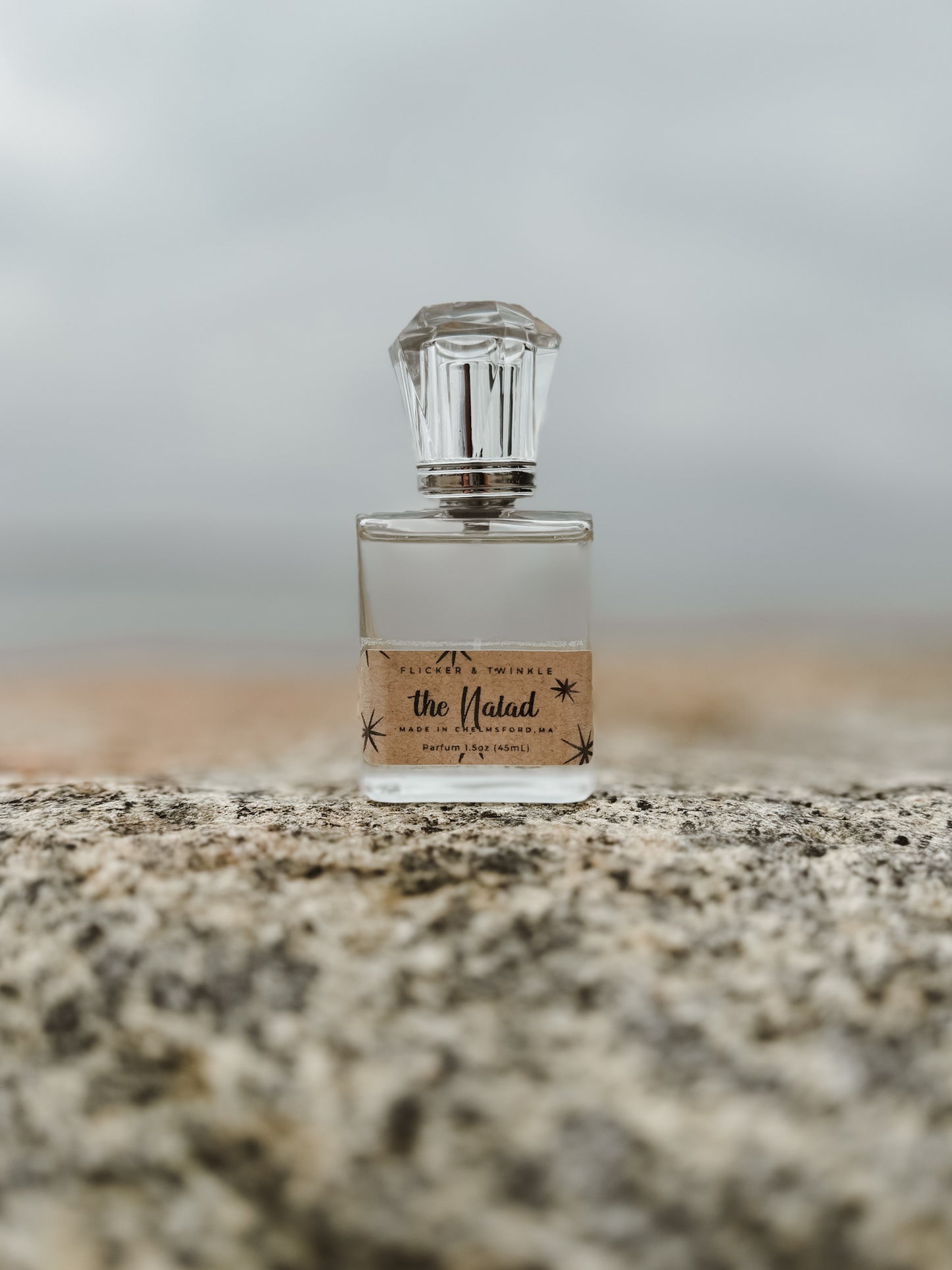 A clear glass bottle of perfume with a crystal-like cap is placed on a textured stone surface. The label on the bottle reads "The Naiad" with decorative black stars and cursive script under the brand name "Flicker And Twinkle." Evoking coastal elegance, the background is blurry, featuring a soft, muted landscape reminiscent of ocean air.