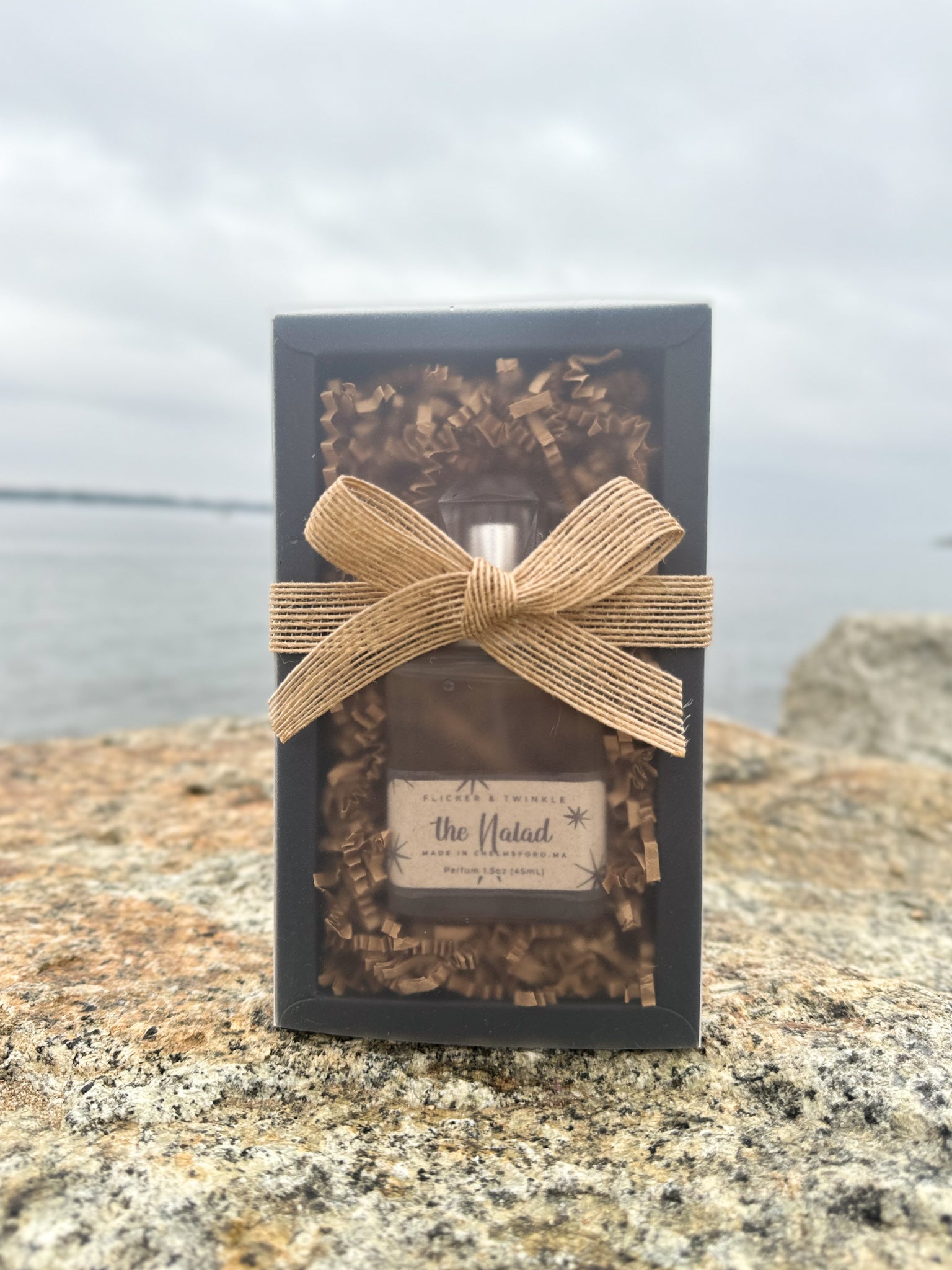 A bottle of "The Naiad" cologne by Flicker And Twinkle is elegantly packaged in a black box with a clear front, accentuated by decorative paper inside. The coastal elegance of the box is highlighted by a tan burlap ribbon. Placed on a rocky surface near water under a cloudy sky, the packaging evokes the scent of ocean air mingled with driftwood.