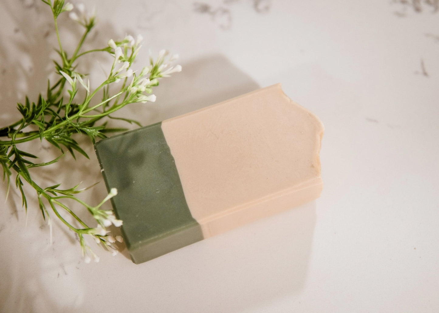 A rectangular goat milk soap bar in sage green and beige rests on a white marble surface. Small white flowers and green foliage lie next to it. The soap has a corner bitten off.