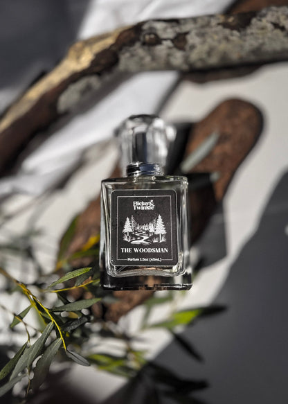 A square glass perfume bottle labeled 'The Woodsman' by 'Flicker and Twinkle' is centered against a styled background of white fabric, dark wood branches, and green leaves. The bottle is clear glass with a black label and a silver-toned cap. The scent is a woodsy Oakmoss and Amber fragrance