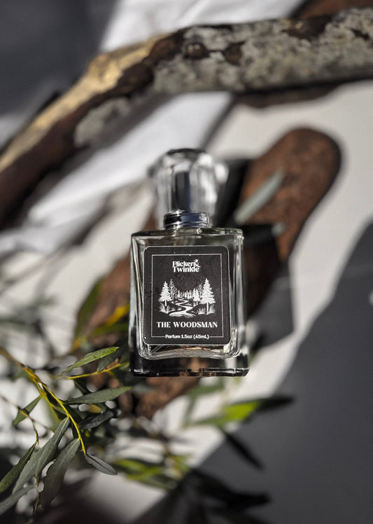 A square glass perfume bottle labeled 'The Woodsman' by 'Flicker and Twinkle' is centered against a styled background of white fabric, dark wood branches, and green leaves. The bottle is clear glass with a black label and a silver-toned cap. The scent is a woodsy Oakmoss and Amber fragrance