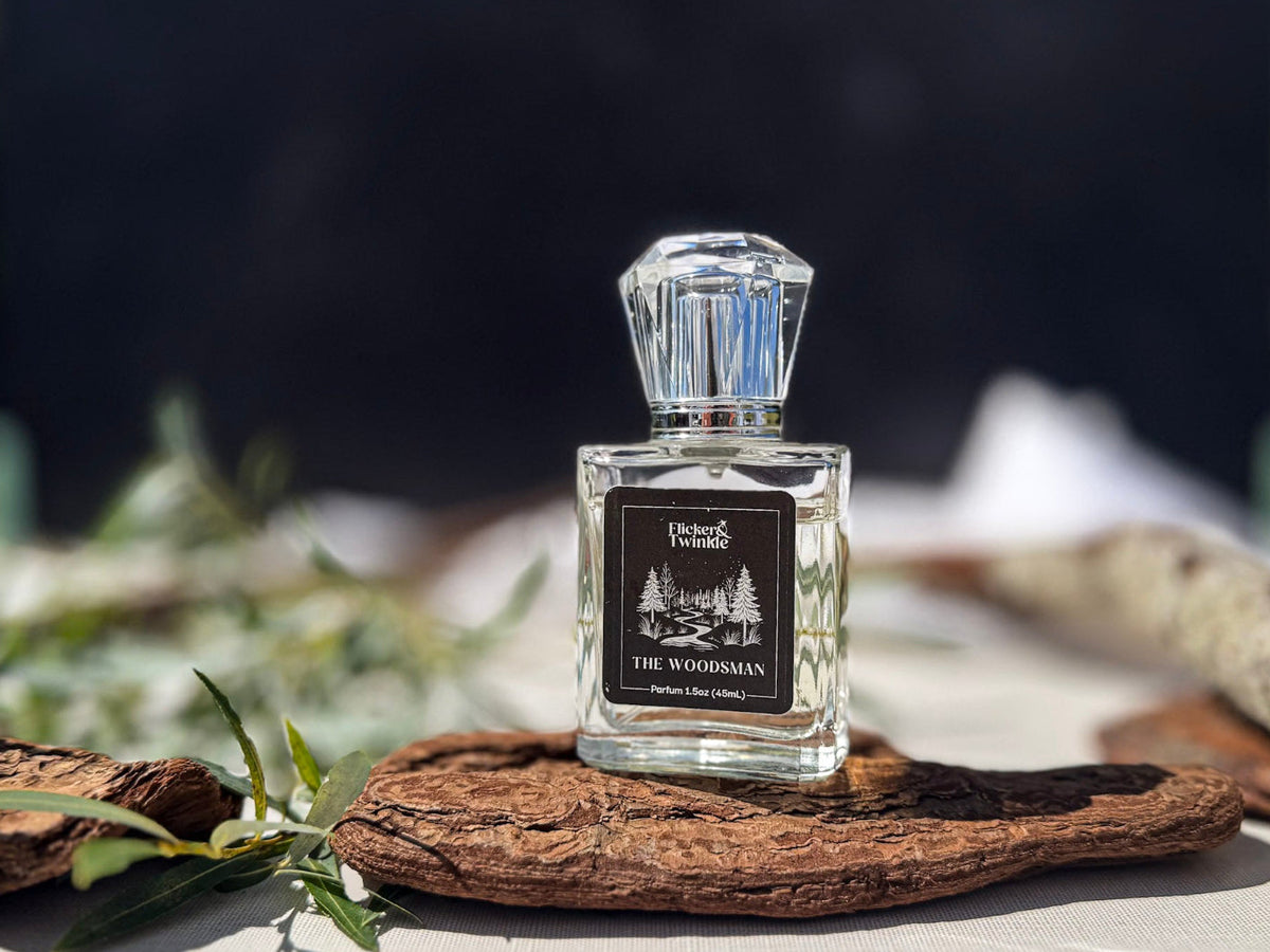 A clear glass perfume bottle with a faceted cap and a black label reading 'The Woodsman' by 'Flicker and Twinkle,' styled on a piece of driftwood outdoors.