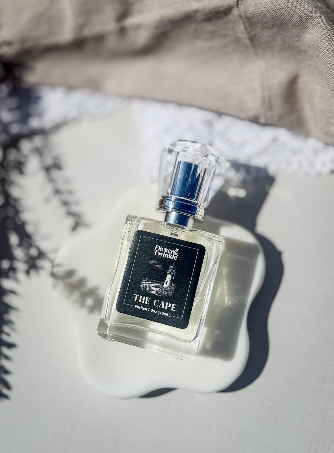 Clear perfume bottle with black and white label on a white flower shaped coaster with an off-white and linen background. The Perfume label brand is Flicker and Twinkle and the name of the scent is "The Cape" and there is a white line drawing of a lighthouse and the sea in the middle of the label.