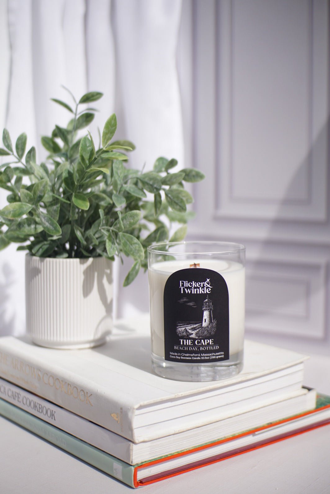 Coconut Soy Beeswax candle in a glass container sits on stacked books beside a small plant, .