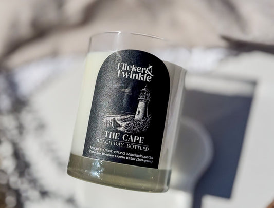 A Flicker & Twinkle 'The Cape' scented candle in a clear glass jar, featuring a black label with a lighthouse illustration, set against a softly blurred background.