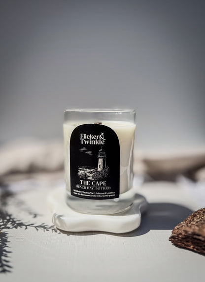 A candle in a clear glass jar with a black label reading 'Flicker & Twinkle The Cape,' placed on a white stone coaster with a softly blurred neutral background. Beach scented Coconut Soy Beeswax Candle