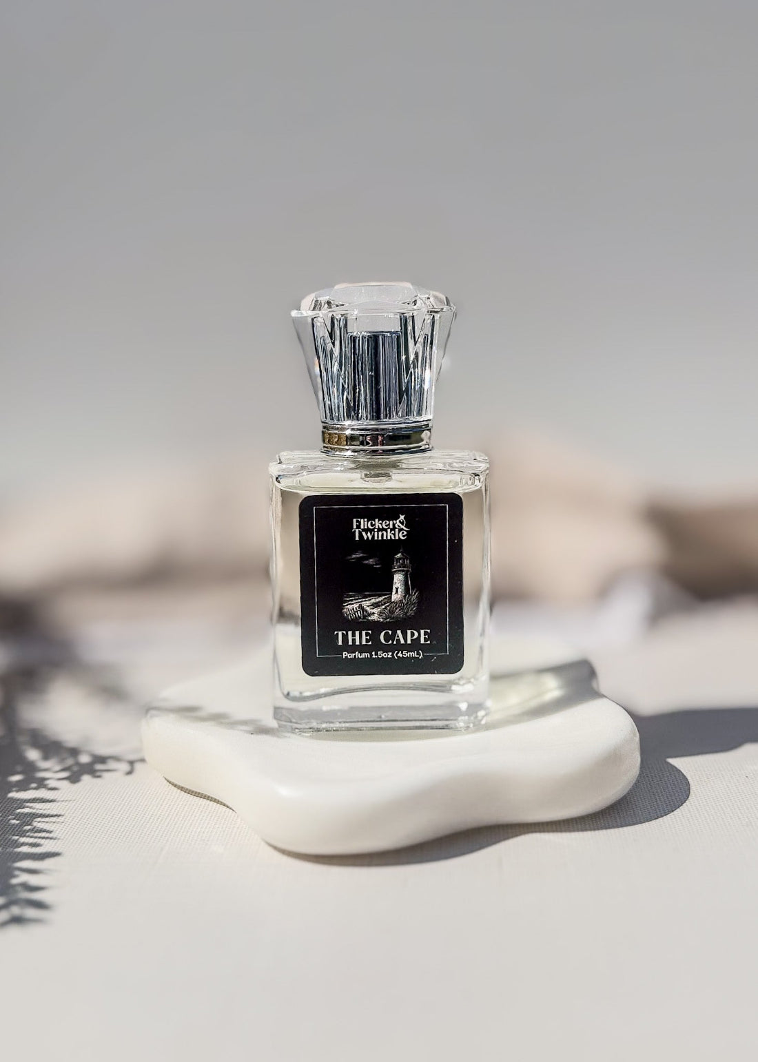 The Cape Perfume Bottle, product photo in front of a creamy off white background. The label is black and white and has an image of a lighthouse on the bottlle. This is a beach inspired perfume scent.