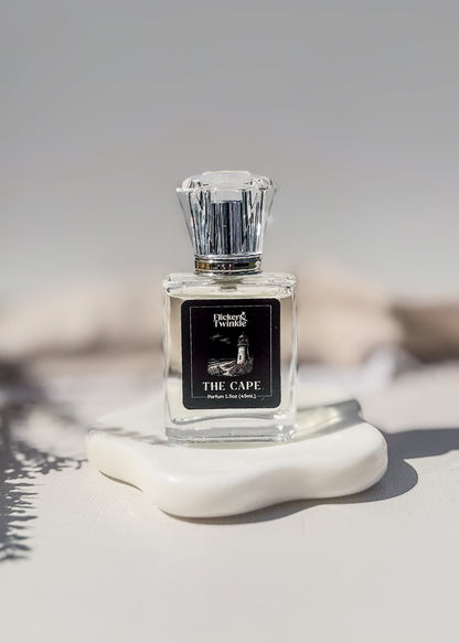 The Cape Perfume Bottle, product photo in front of a creamy off white background. The label is black and white and has an image of a lighthouse on the bottlle. This is a beach inspired perfume scent.