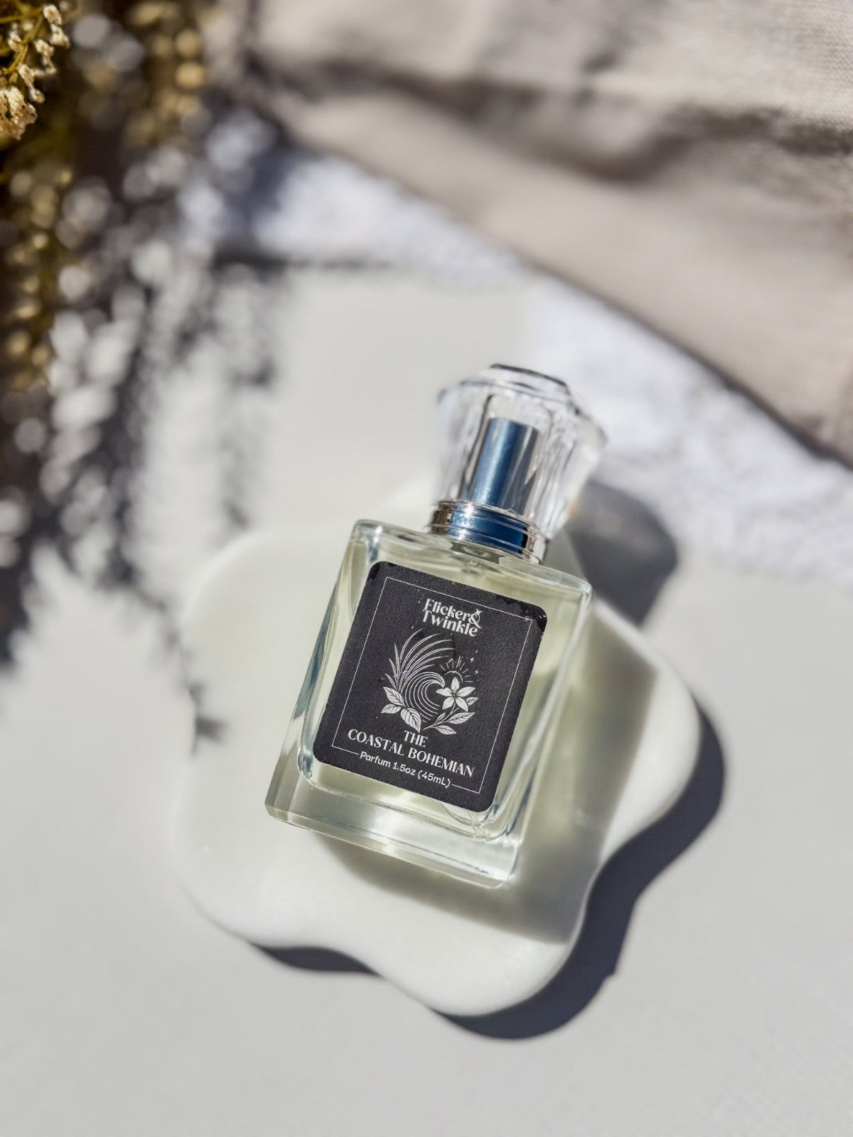 A square glass perfume bottle with a clear cap and a black label reading 'The Coastal Bohemian', styled on a white ceramic dish against a light gray background with soft shadows. The Coastal Bohemian is a blend of Sea Salt Neroli and Patchouli.