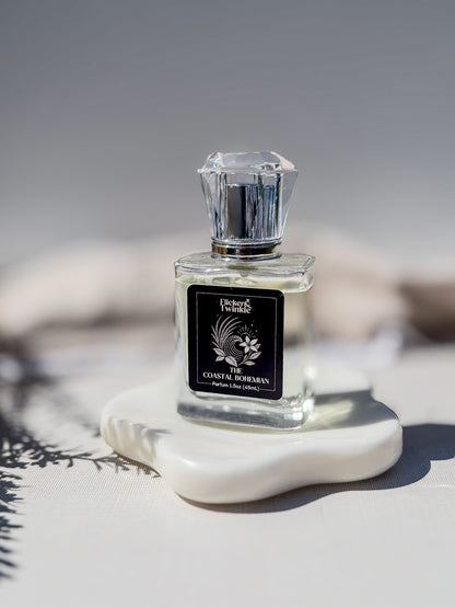 A clear glass perfume bottle with a faceted silver cap and a black label reading The Coastal Bohemian, displayed on a white ceramic dish against a soft gray background.
