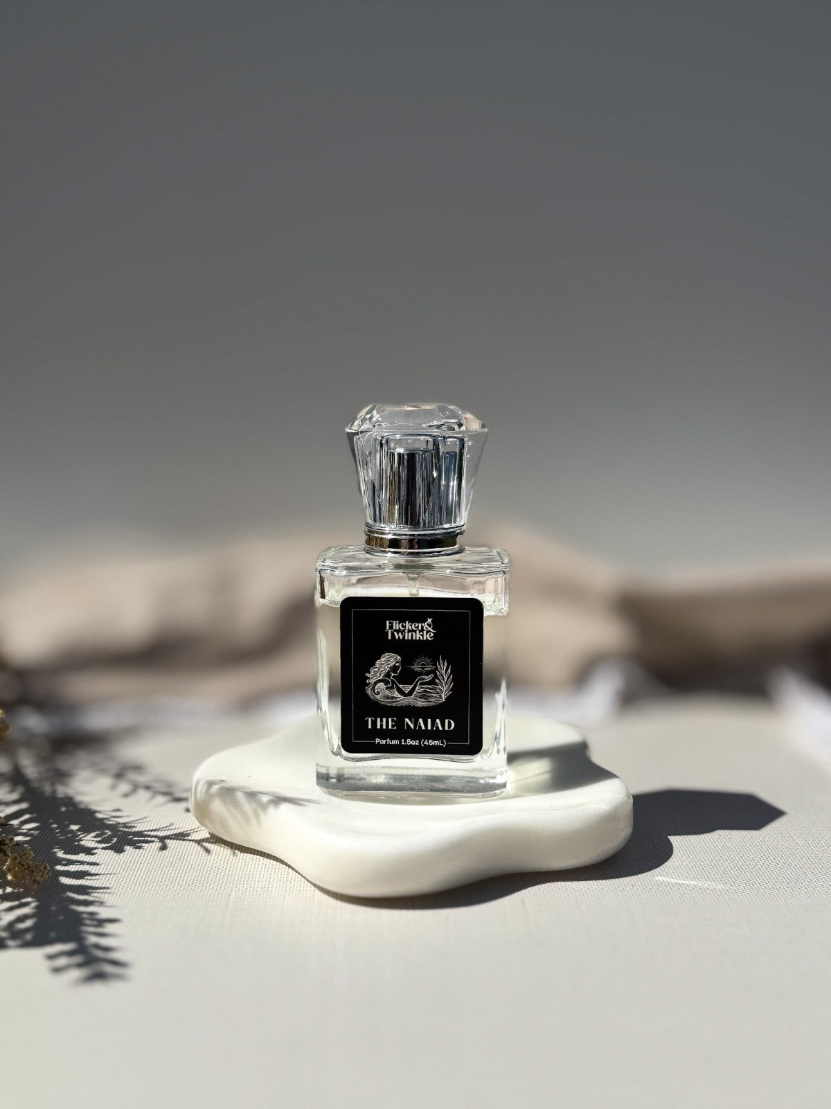 A clear glass perfume bottle with a faceted clear cap and a black front label reading 'The Naiad" displayed on a white sculptural base against a soft gray background. The Naiad is a clean aquatic scent with some white florals to balance.