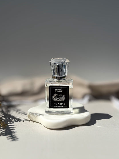 A clear glass perfume bottle with a faceted clear cap and a black front label reading 'The Naiad" displayed on a white sculptural base against a soft gray background. The Naiad is a clean aquatic scent with some white florals to balance.