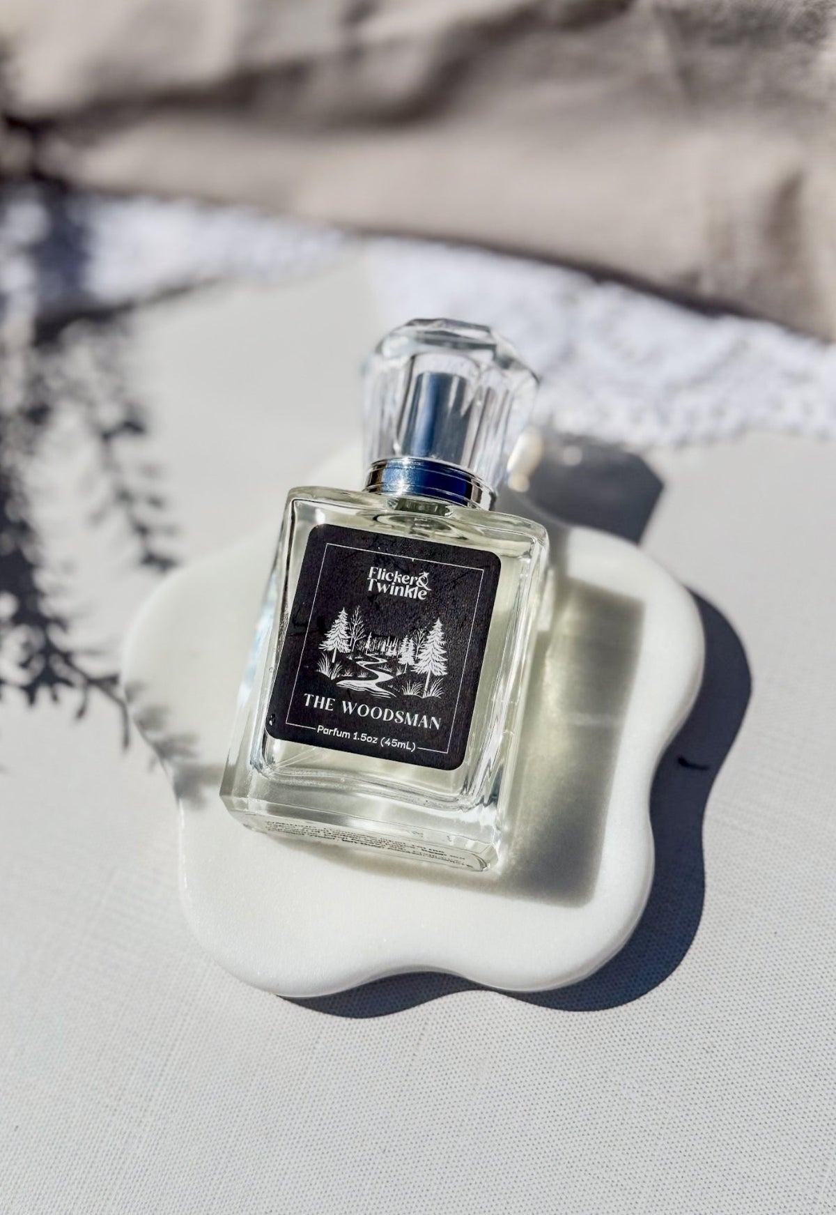 A clear glass perfume bottle labeled 'The Woodsman' by Flicker and Twinkle, placed on a white ceramic dish against a light fabric background with soft shadows.