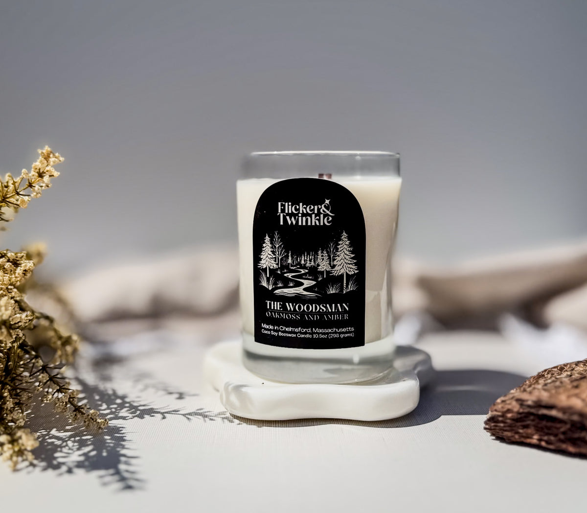 A Flicker & Twinkle coconut soy beeswax soy wood wick candle in a clear glass jar with a black label, displayed on a white stone coaster with dried flowers and a wooden slice in the foreground.