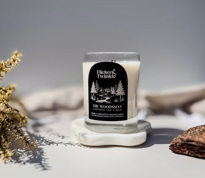 A Flicker & Twinkle coconut soy beeswax soy wood wick candle in a clear glass jar with a black label, displayed on a white stone coaster with dried flowers and a wooden slice in the foreground.