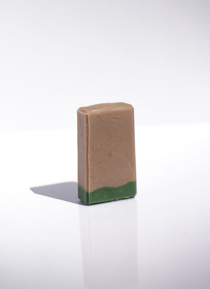 The Woodsman Goats Milk Soap, a green rectangular bar, stands upright on a white surface.