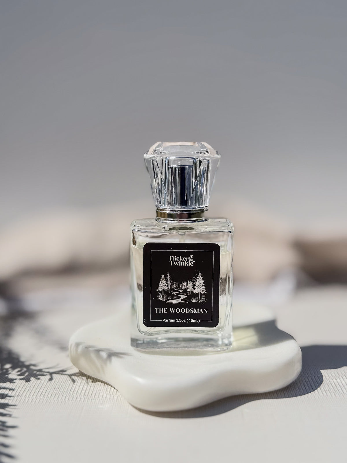 A clear glass perfume bottle with a silver faceted cap and a black label reading 'Flicker and Twinkle' and 'THE WOODSMAN', displayed on a white sculptural stand against a soft gray background.