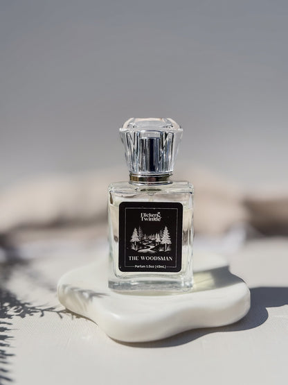 A clear glass perfume bottle with a silver faceted cap and a black label reading 'Flicker and Twinkle' and 'THE WOODSMAN', displayed on a white sculptural stand against a soft gray background.