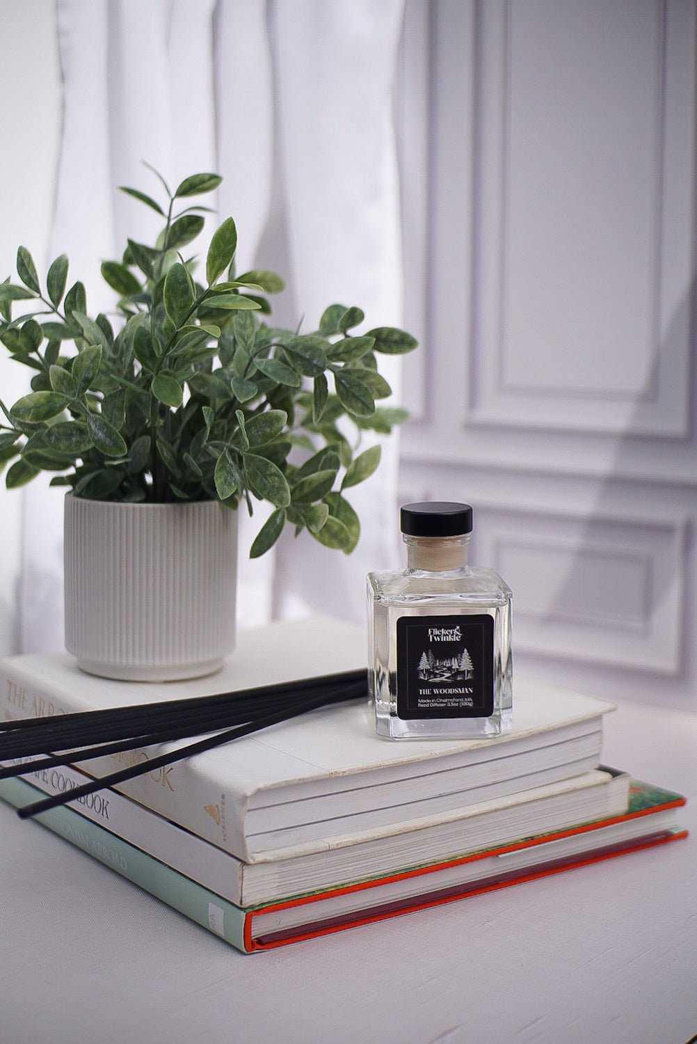 A potted plant, The Woodsman Reed Diffuser, and books on a white surface near a window.
