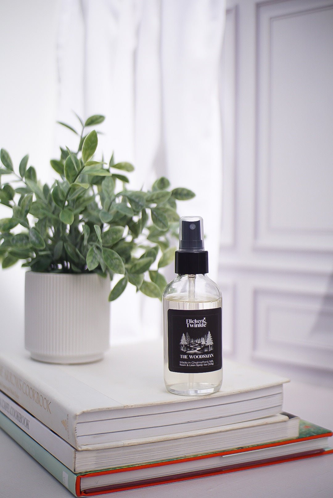 The Woodsman Room and Linen Spray on stacked books beside a potted plant on a white surface.