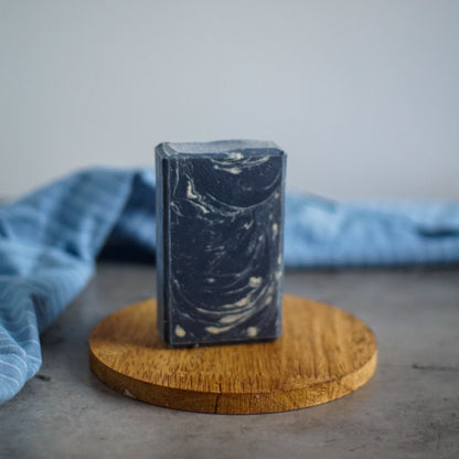 The Yachtsman Goat Milk Soap by Flicker And Twinkle, a rectangular marbled bar in dark grey and white hues, stands upright on a round wooden coaster. The backdrop of blue fabric gently draped over a grey surface evokes an oceanic scent that refreshes the senses.