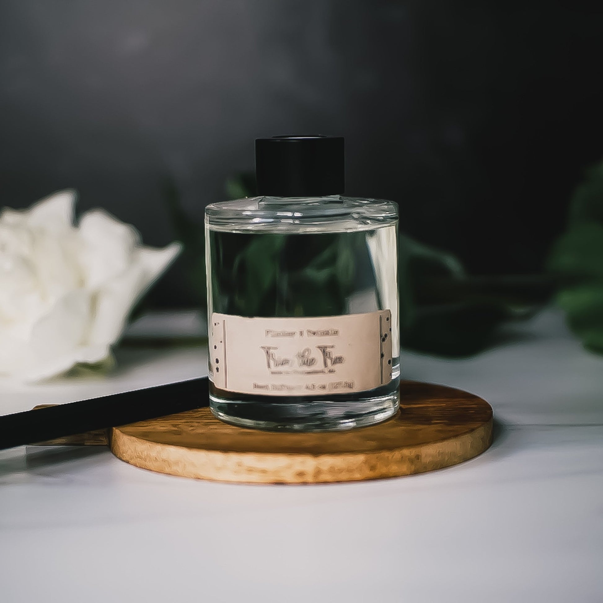 A clear, rectangular bottle with a black cap sits on a round wooden base. The bottle features a label on the front and contains the Trim the Tree Reed Diffuser from Flicker And Twinkle. In the background, there is a blurred white flower and green leaves.