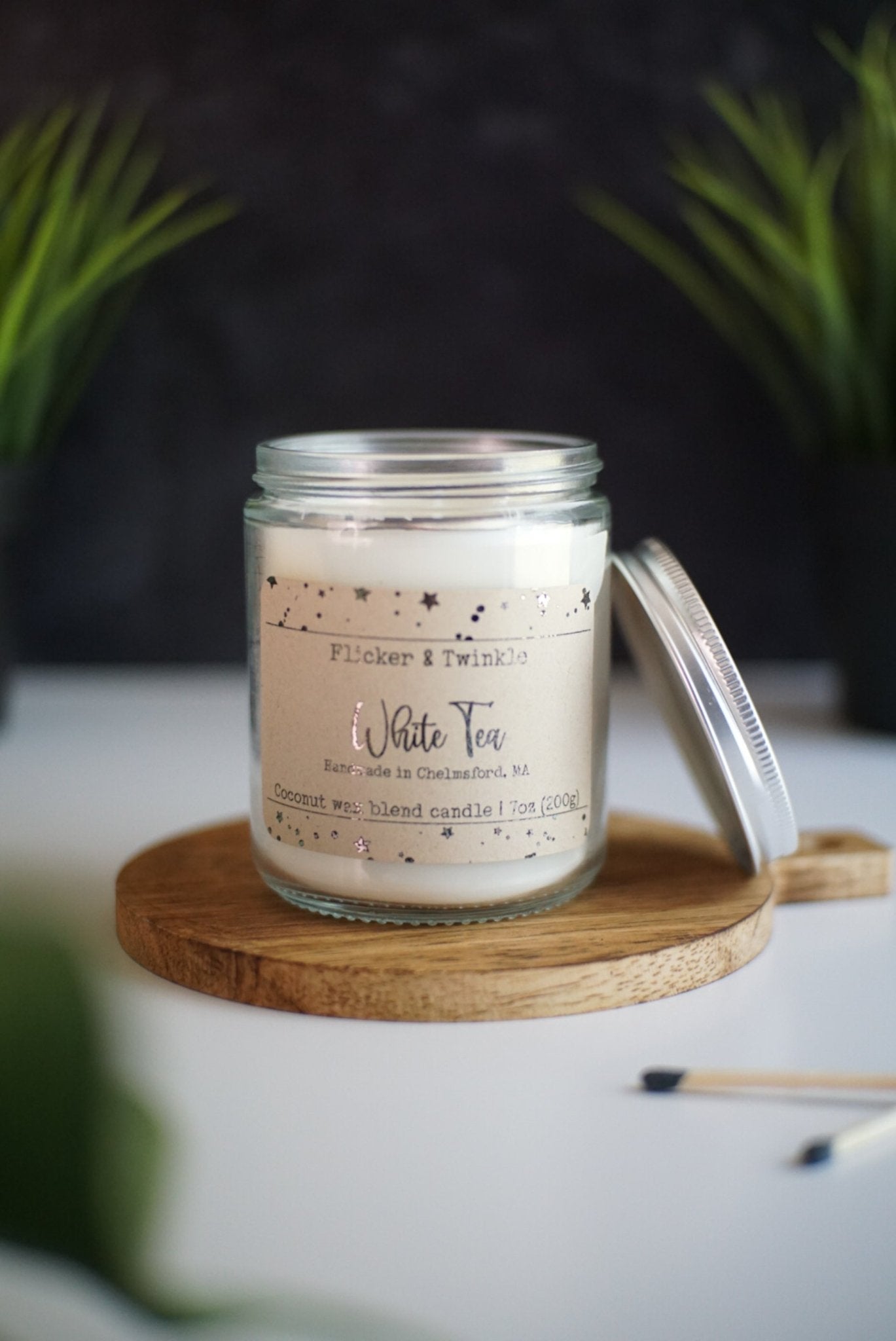 A lit White Tea Coconut Wax Candle by Flicker And Twinkle in a glass jar sits on a wooden coaster. The label reads "Flicker And Twinkle, White Tea, Handmade in Chelmsford, MA, Coconut wax blend candle, 7 oz (200 g)." The jar lid is propped against the jar, with two matchsticks visible in the foreground.