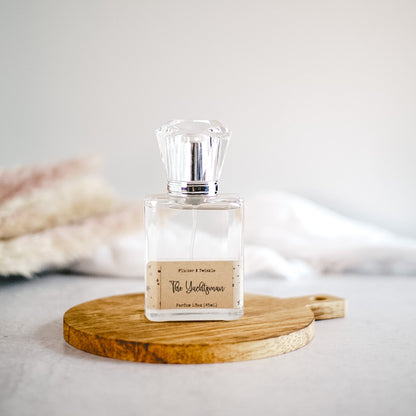 A clear perfume bottle with a faceted cap and a label reading "The Gentlewoman" is placed on a small round wooden board. The softly blurred background, rich with neutral tones and textures, captures the oceanic essence reminiscent of "The Yachtsman" fragrance by Flicker And Twinkle.