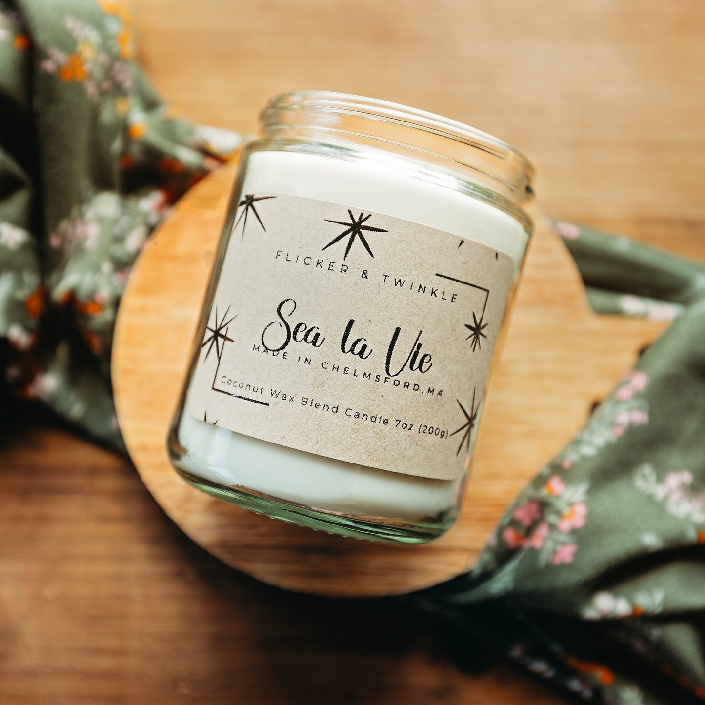 A jar candle labeled "Flicker And Twinkle - Sea La Vie Coconut Wax Candle - Made in Chelmsford, MA - 7oz (200g)" is displayed on a wooden surface. Emitting an ocean scent that promises relaxation and serenity, the candle is nestled next to green floral-patterned fabric draped around it.