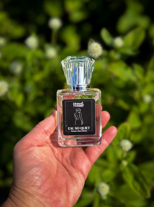 A hand holding a small clear perfume bottle labeled 'Eau So Quiet' by the brand 'Flicker and Twinkle' against a blurred green leafy background.
