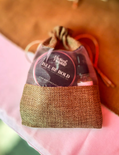 A small burlap drawstring pouch containing a round car freshener tin labeled 'ISLE BE BOLD' and a small glass refill bottle, resting on a white tablecloth with a warm brown background.