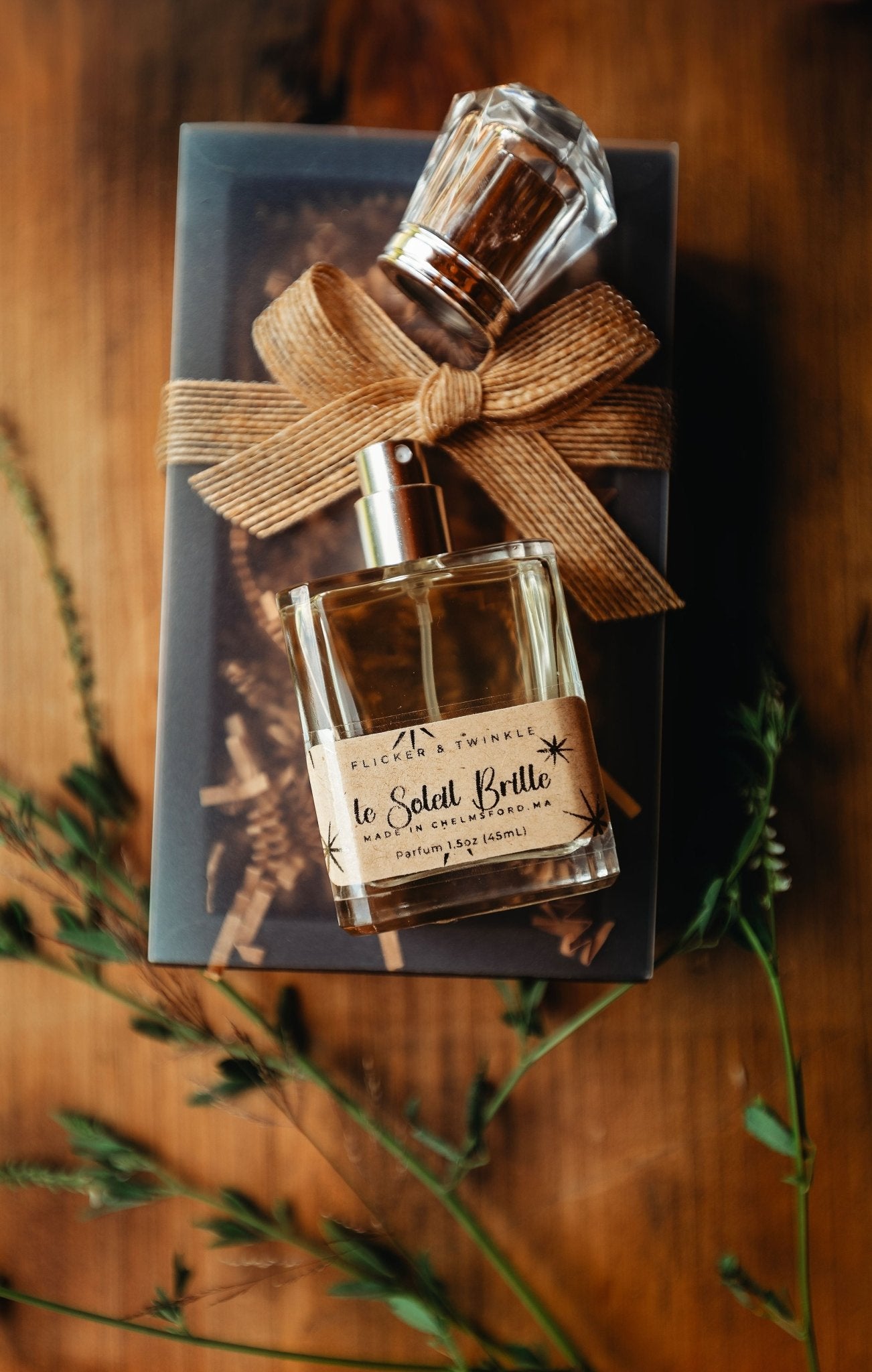 A glass perfume bottle labeled "Le Soleil Brille Perfume" by Flicker And Twinkle, adorned with a decorative cap, is placed on top of a closed book tied with a tan ribbon bow. Sprigs of greenery are arranged on both sides, all set on a wooden surface, creating an elegant and rustic presentation that evokes the essence of a summer fragrance.