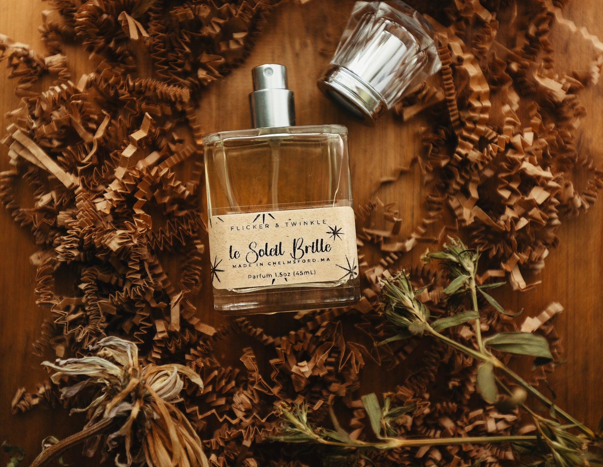 A glass perfume bottle labeled "Flicker And Twinkle: Le Soleil Brille Perfume," surrounded by crinkled paper and dried plants, lies on a wooden surface. The bottle’s lid is placed to the side, hinting at the summer fragrance within, with notes of sandalwood lingering in the air.