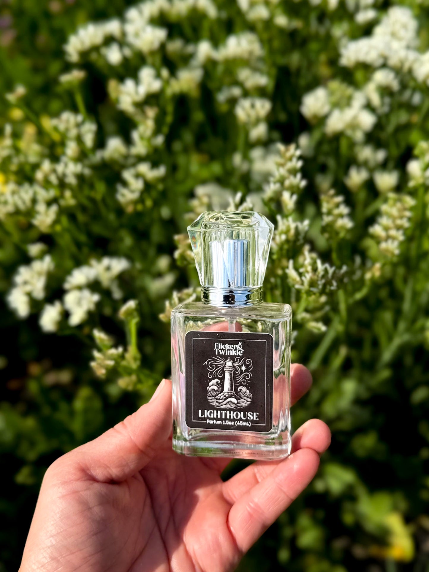 A hand holds a small clear glass perfume bottle labeled 'Lighthouse' by Flicker and Twinkle. against a blurred background of green foliage and white flowers.