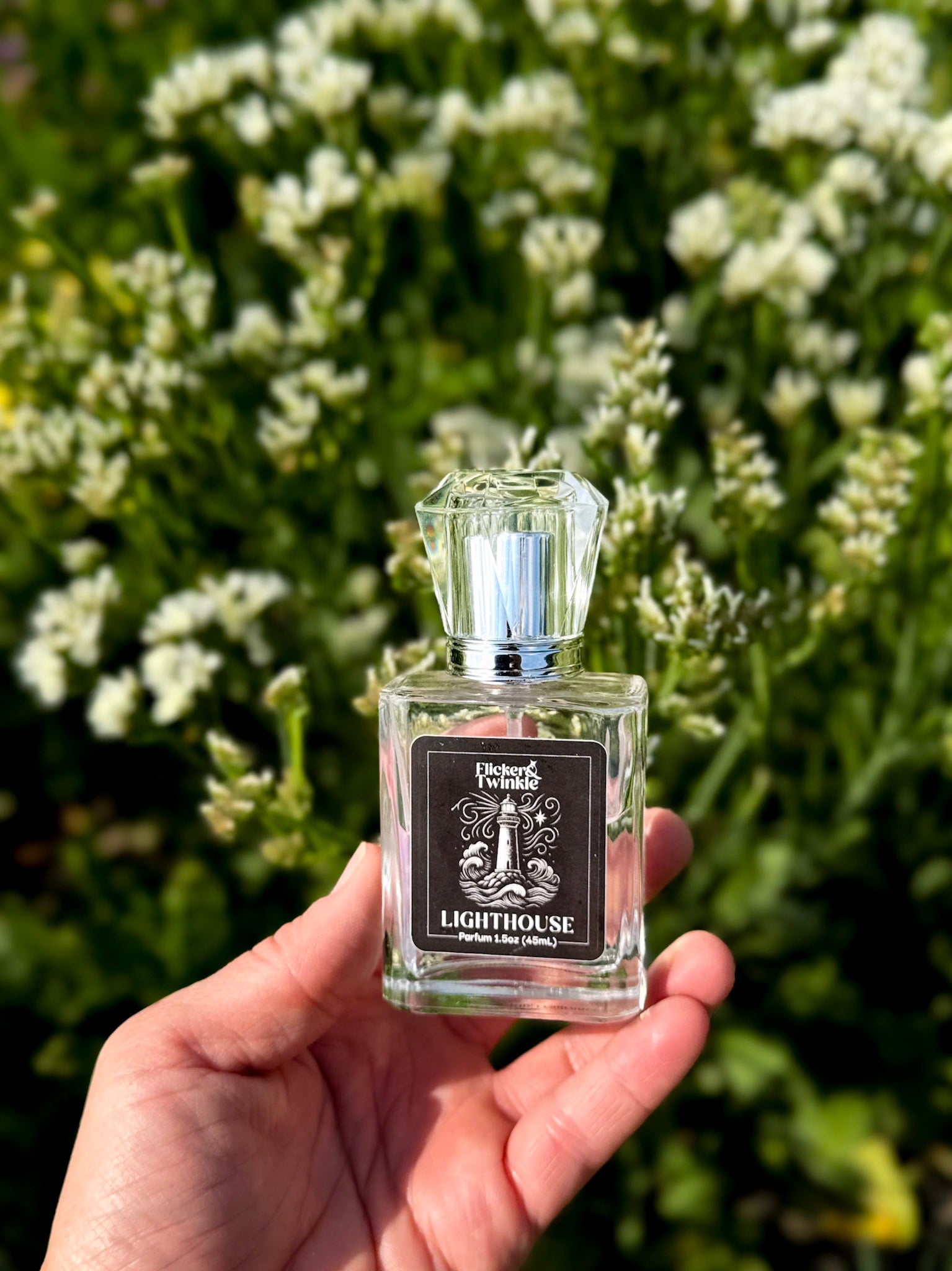 A hand holds a small clear glass perfume bottle labeled 'Lighthouse' by Flicker and Twinkle. against a blurred background of green foliage and white flowers.