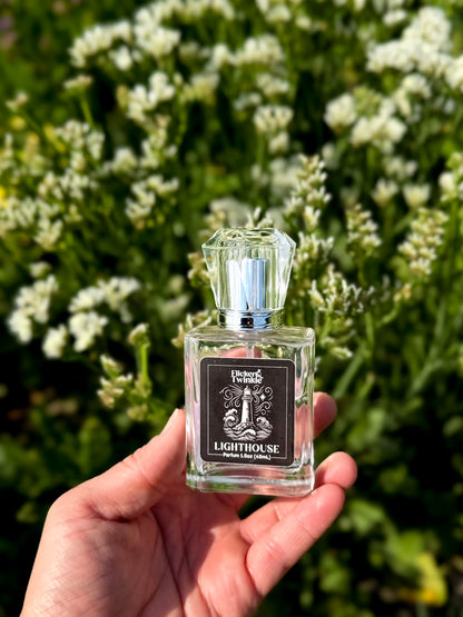 A hand holds a small clear glass perfume bottle labeled 'Lighthouse' by Flicker and Twinkle. against a blurred background of green foliage and white flowers.
