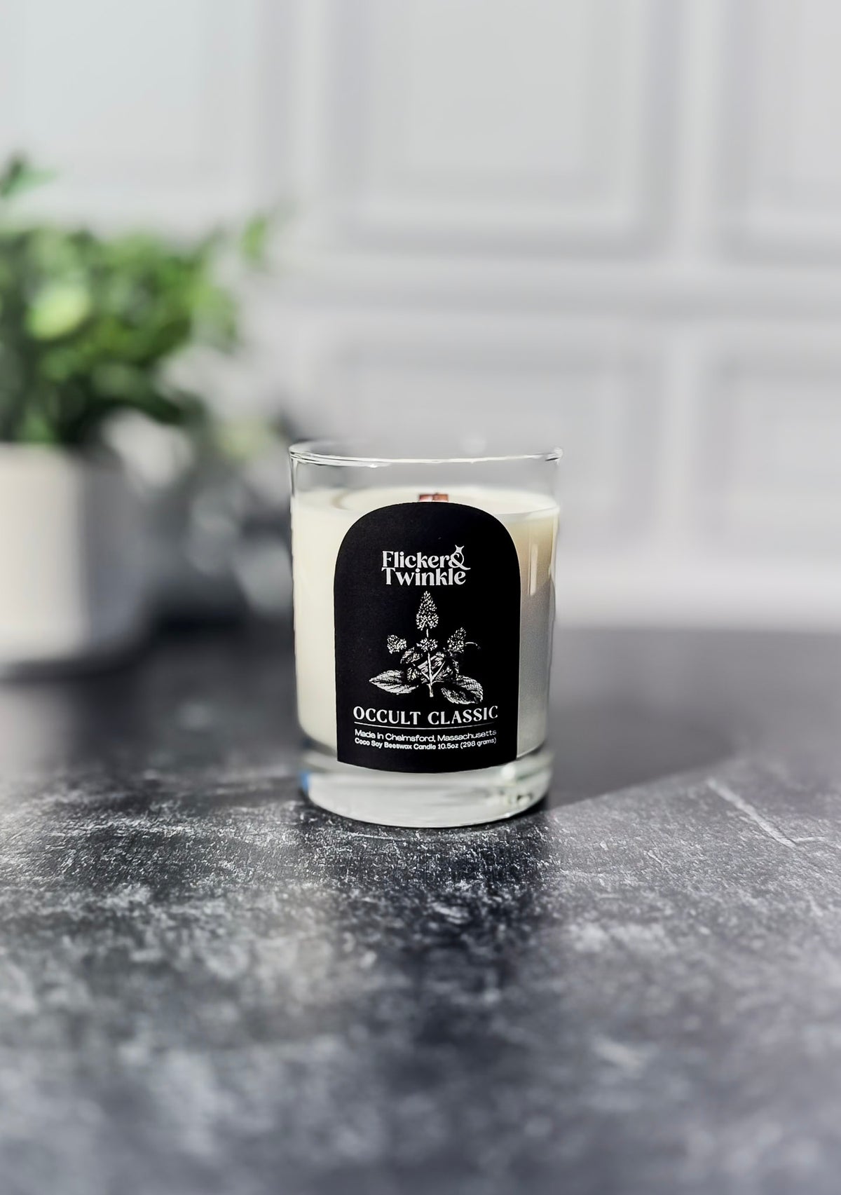 A Fickler & Twinkie Occult Classic scented candle in a clear glass jar, set on a dark gray stone surface with a blurred white paneled wall and green plant in the background.
