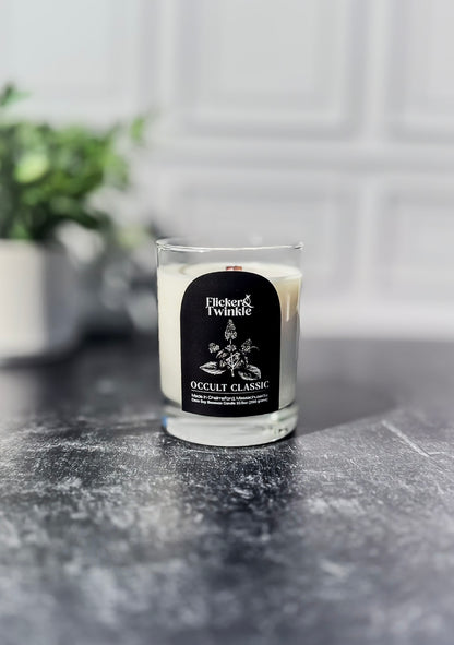A Fickler & Twinkie Occult Classic scented candle in a clear glass jar, set on a dark gray stone surface with a blurred white paneled wall and green plant in the background.