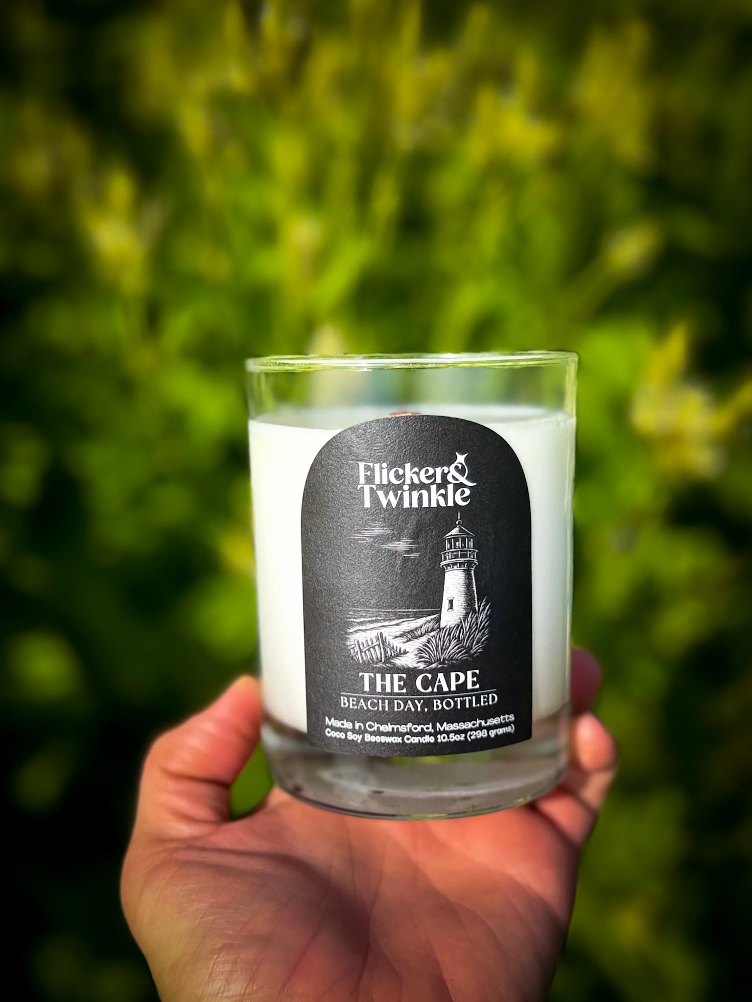 A hand holds a clear glass jar candle with white wax and a black label reading 'Flicker Twinkle' and 'The Cape Beach Day  against a blurred green outdoor background.