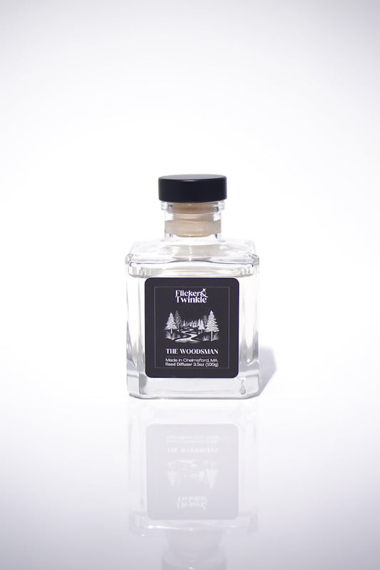 Square glass bottle labeled The Woodsman Reed Diffuser, with black cap, oakmoss and amber woods.