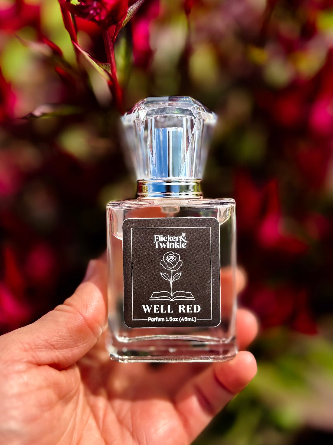 A hand holds a small clear glass perfume bottle with a black label called "Well Red' against a blurred background of dark red foliage. by the company Flicker and Twinkle.